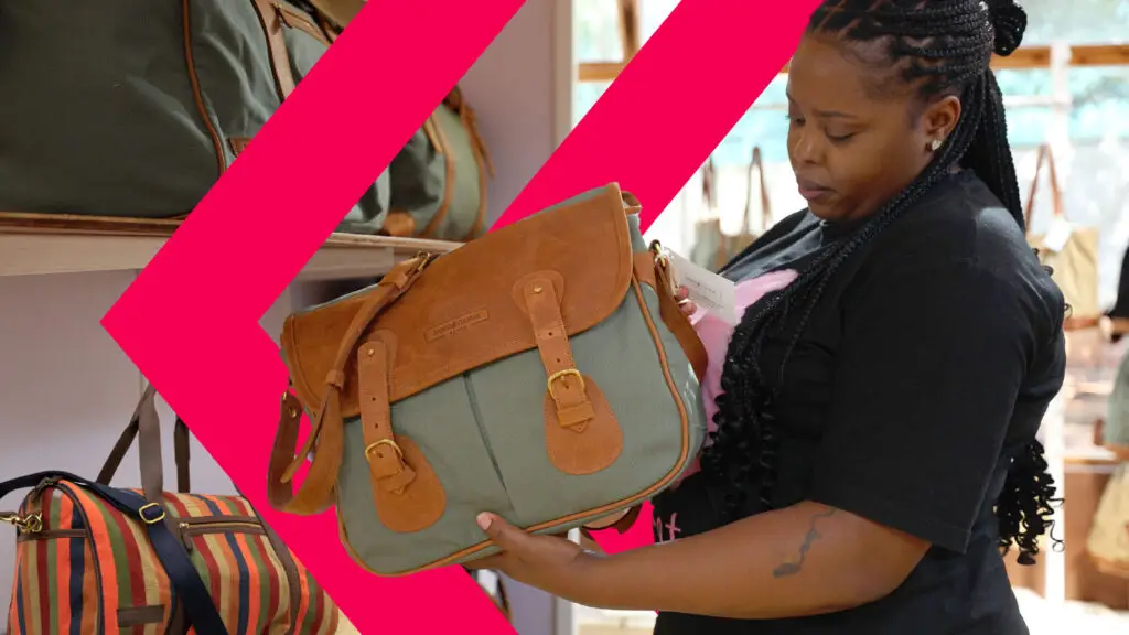 Close-up of a green and tan leather Sandstorm Kenya bag being held in a retail setting, with other luxury bags displayed on shelves in the background.